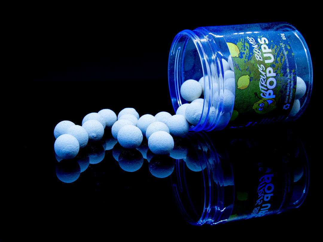 Auf diesem Bild sieht man die weißen Blackdeere Citrus Bomb Pop Ups, welche aus einer runden, offenen, auf dem Boden liegenden Plastikverpackung raus rollen. Der Hintergrund ist dunkel und die Köder sind von einer Schwarzlichtlampe angeleuchtet und zeigen Ihren fluorisierenden Charakter.